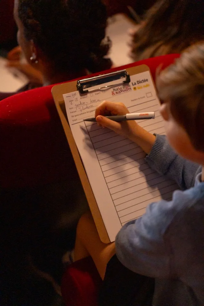 Un enfant remplit le formulaire de la dictée culinaire, posé sur une planchette, dans un fauteuil de cinéma rouge